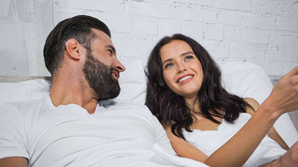 happy brunette woman talking to smiling boyfriend while lying in bed - Photo, Image