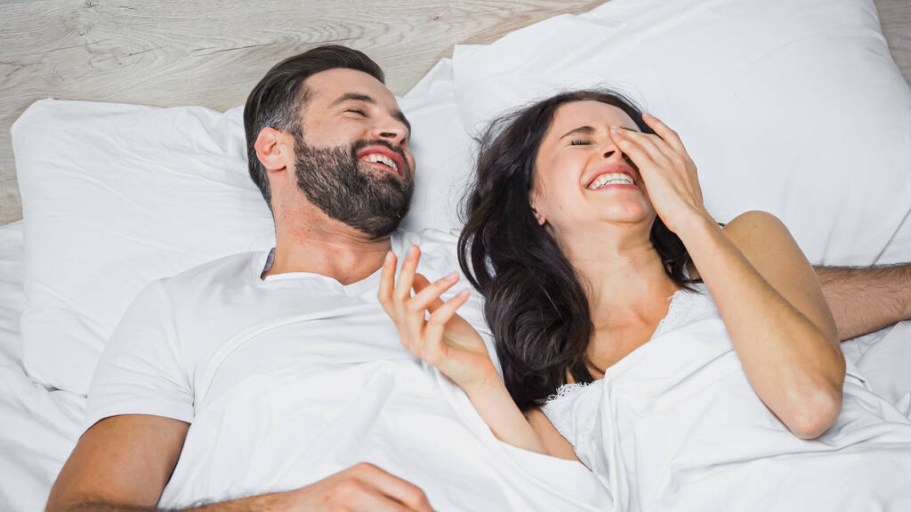 смеющаяся женщина трогает лицо, лежа в постели рядом с счастливым мужчиной - Фото, изображение