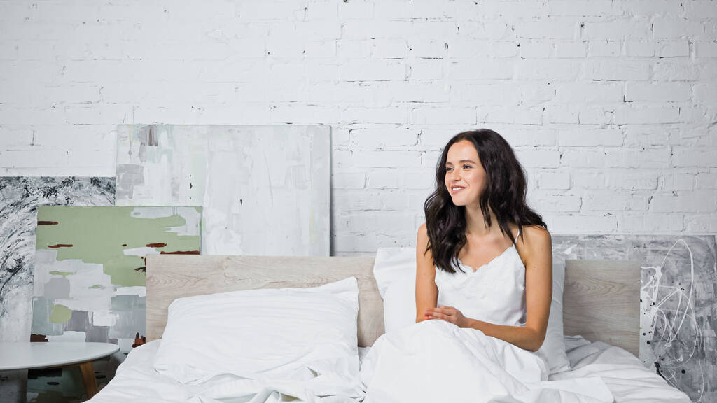 fröhliche brünette Frau lächelt, während sie morgens auf dem Bett sitzt - Foto, Bild