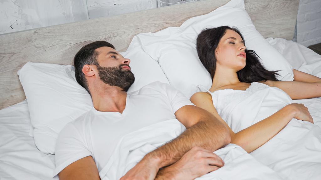 γενειοφόρος άντρας κοιτάζει μελαχρινή γυναίκα κοιμάται κοντά στο κρεβάτι - Φωτογραφία, εικόνα