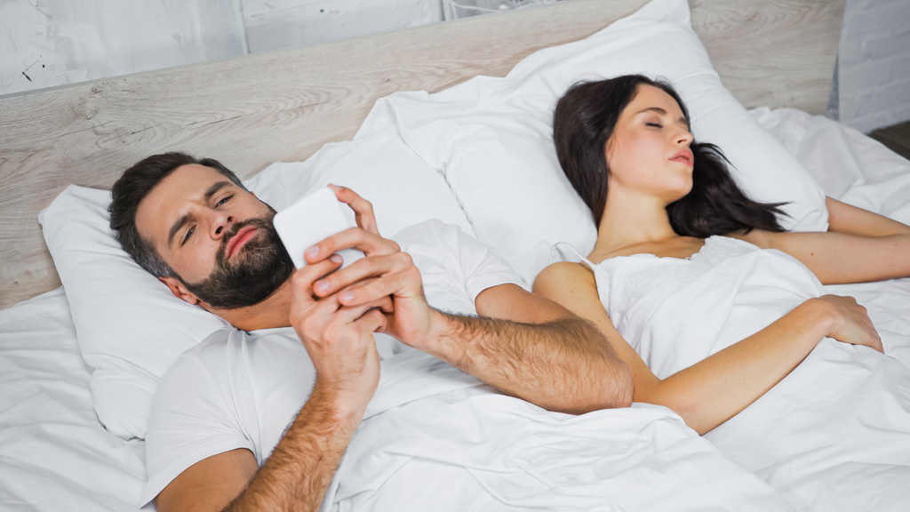 Sakallı adam cep telefonuyla konuşuyor. Yatakta uyuyan kız arkadaşının yanında. - Fotoğraf, Görsel