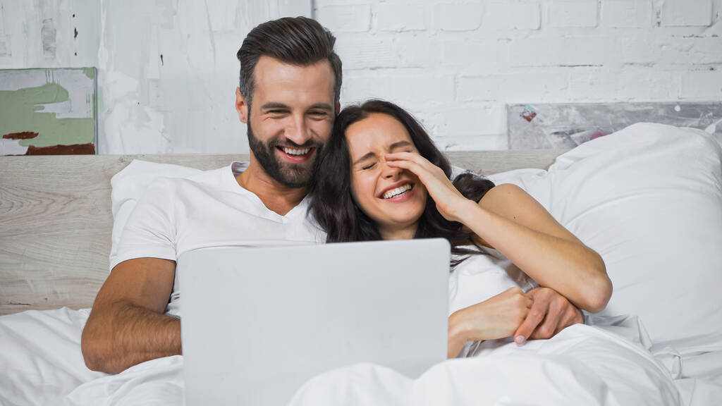 ベッドで陽気な彼氏とノートパソコンで映画を見ながら顔に触れる笑っている女性 - 写真・画像