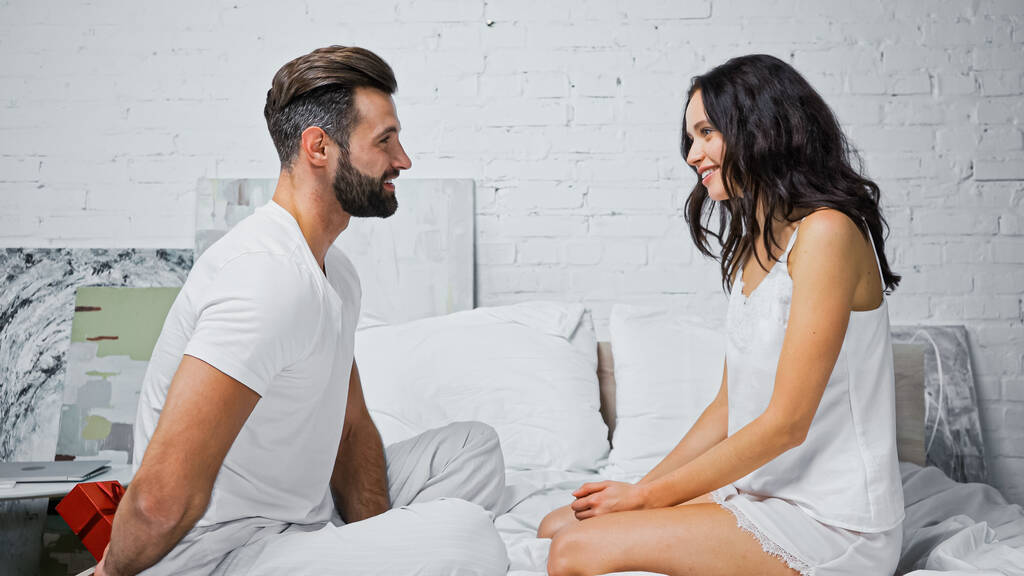 side view of man holding gift box behind back while sitting on bed near smiling girlfriend - Photo, Image