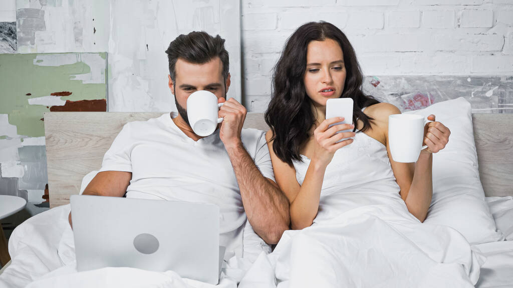 νεαρό ζευγάρι πίνοντας καφέ και χρησιμοποιώντας gadgets στην κρεβατοκάμαρα - Φωτογραφία, εικόνα