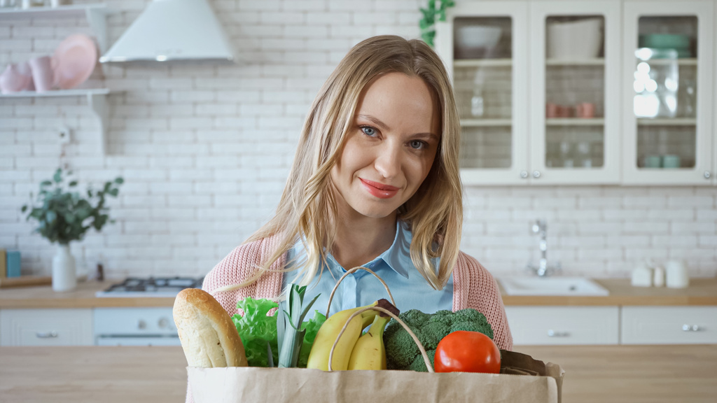 Mujer sonriente con verduras frescas y baguette en bolsa de papel en la cocina  - Foto, imagen