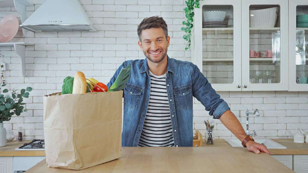 Homme souriant regardant la caméra près de la nourriture fraîche dans la cuisine  - Photo, image