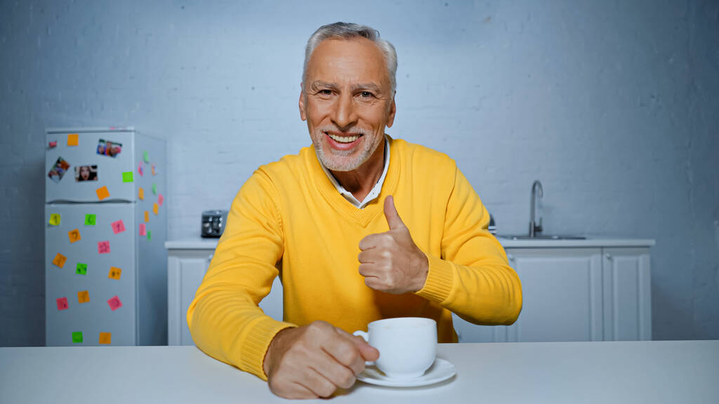 Usmívající se starší muž ukazuje jako blízký šálek kávy v kuchyni  - Fotografie, Obrázek