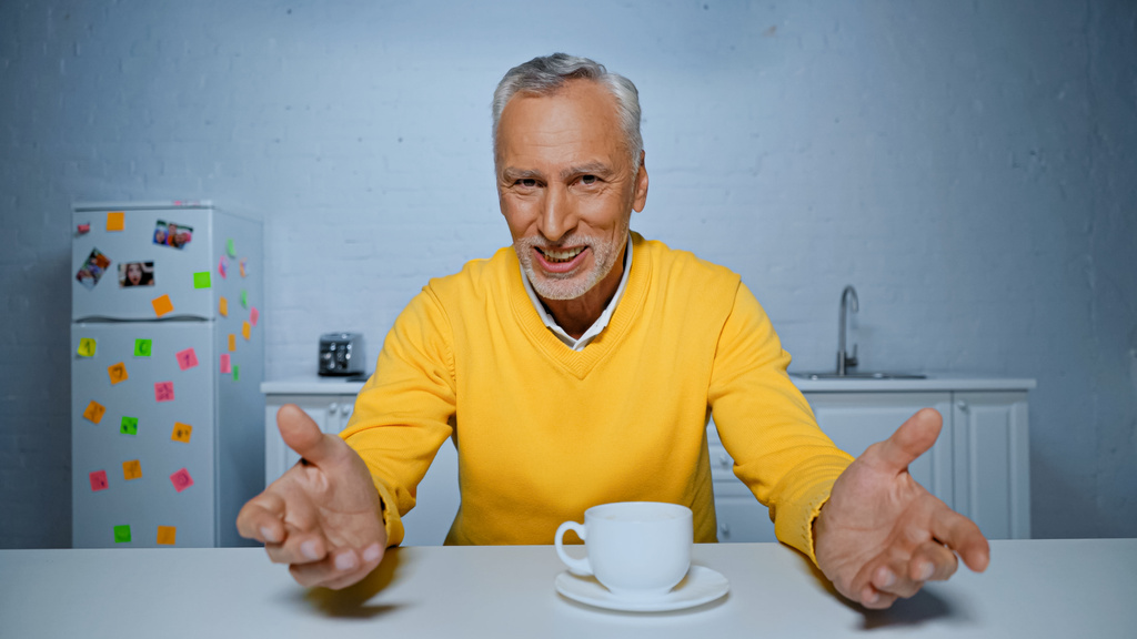 Усміхнений чоловік вказує руками на камеру біля чашки на кухонному столі
  - Фото, зображення