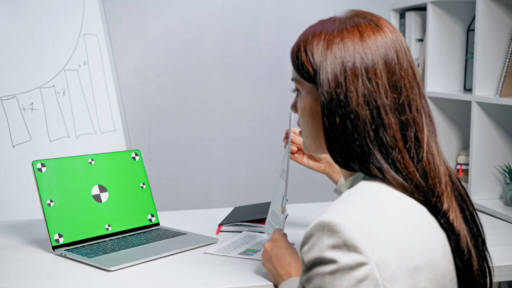 İş kadınının yanında yeşil ekranlı dizüstü bilgisayar ofiste bulanık önplanda belge gösteriyor  - Fotoğraf, Görsel