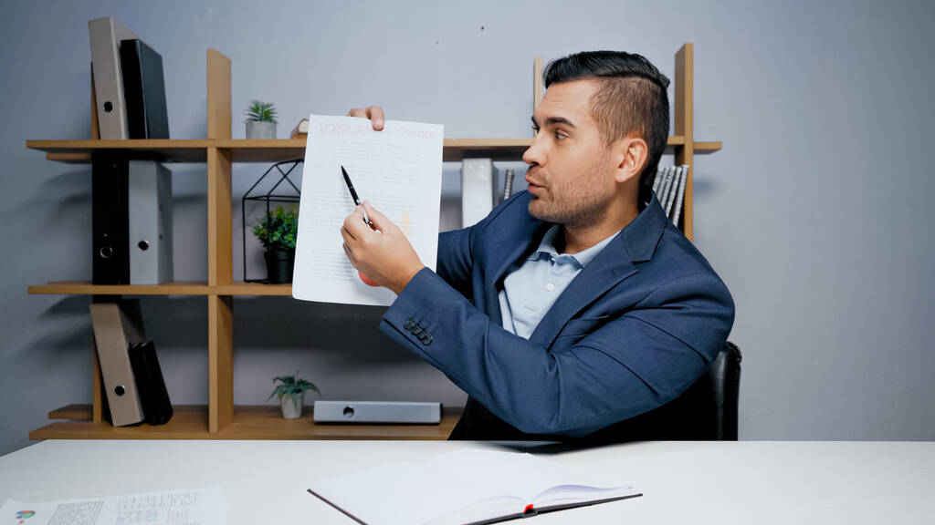 Geschäftsmann zeigt mit Stift auf Papier neben Notizbuch auf Tisch  - Foto, Bild