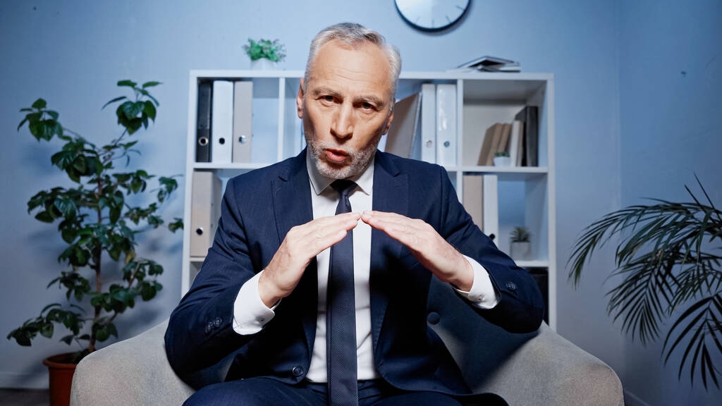 Grijsharige zakenman aan het praten en gebaren tijdens videogesprek in kantoor  - Foto, afbeelding