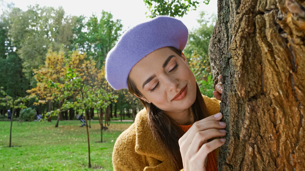 glückliche junge Frau lächelt, während sie Baumstamm im Park berührt - Foto, Bild
