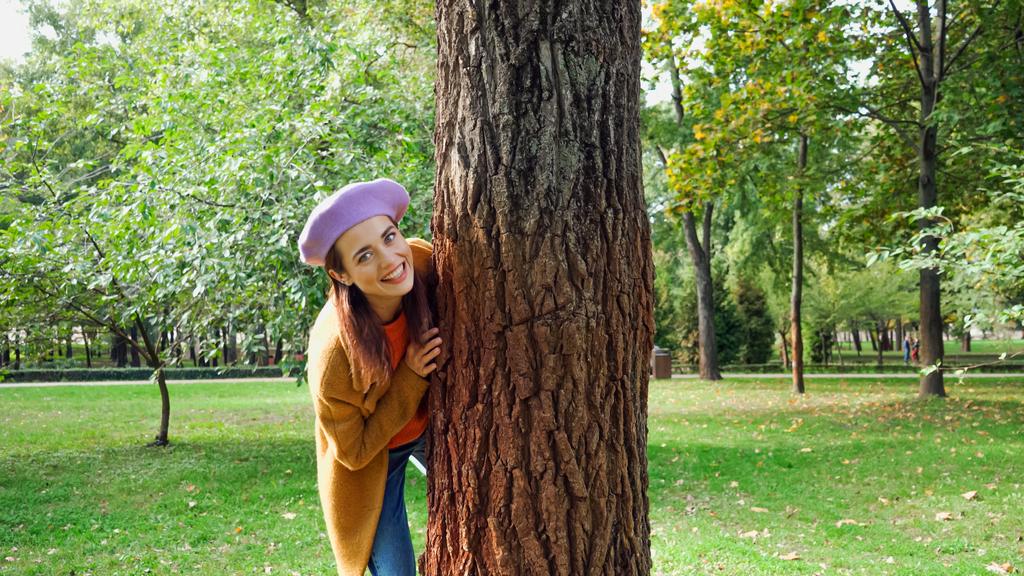 カメラを見ながら公園の木の幹の後ろに隠れている興奮した女性 - 写真・画像