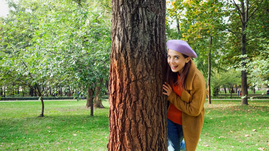 公園の木の幹の後ろに隠れてカメラを見ている興奮した女性 - 写真・画像