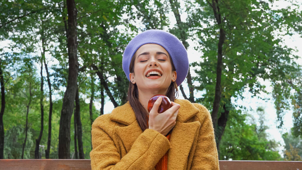 взволнованная женщина смеется с закрытыми глазами, держа спелое яблоко в парке  - Фото, изображение