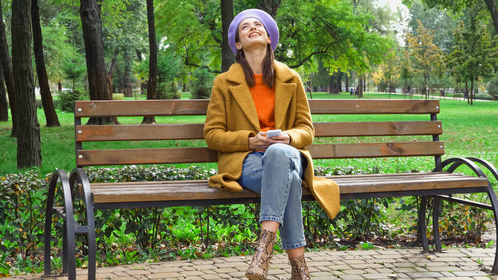 donna allegra guardando in alto mentre messaggistica su smartphone su panchina nel parco - Foto, immagini