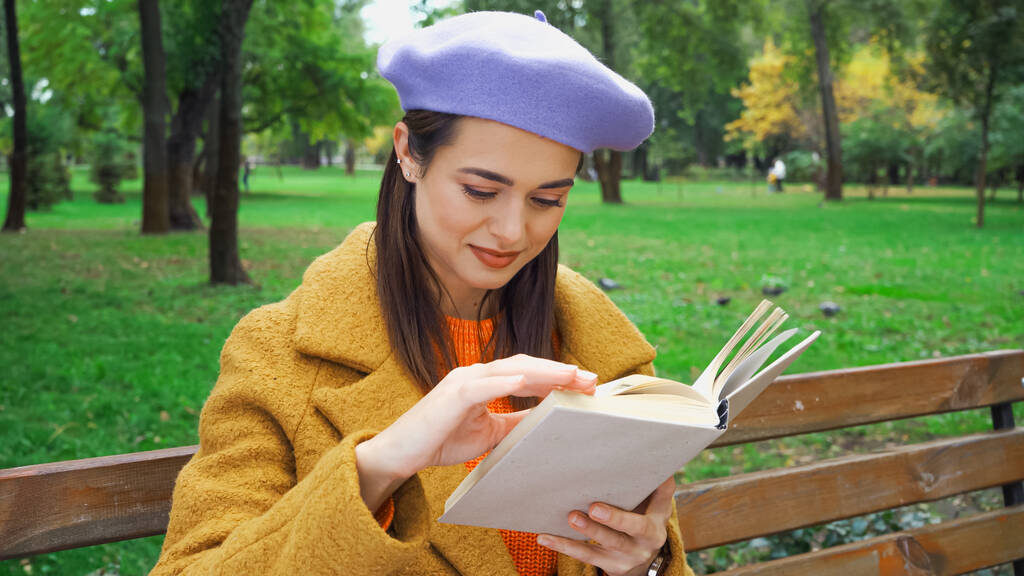 Lächelnde Frau im trendigen Herbst-Outfit liest Buch im Park - Foto, Bild