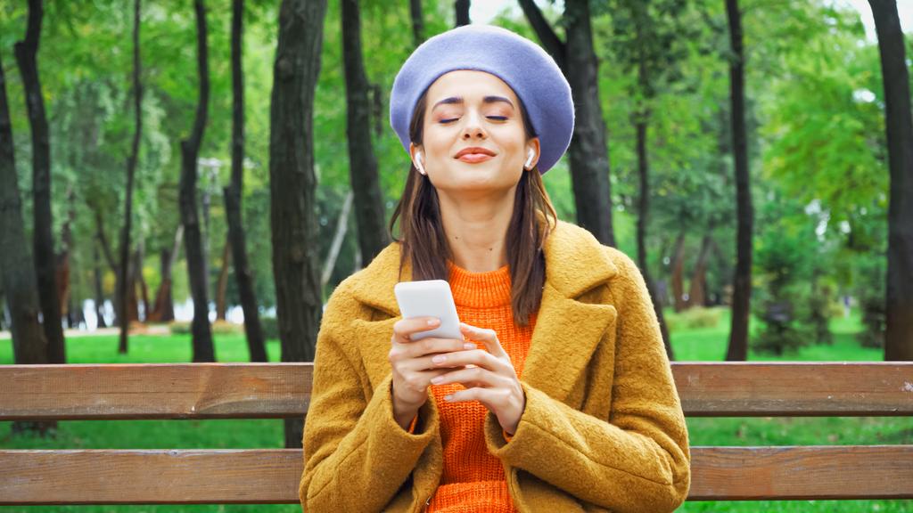 ευχαριστημένη γυναίκα με κομψά φθινοπωρινά ρούχα ακούγοντας μουσική στο πάρκο  - Φωτογραφία, εικόνα
