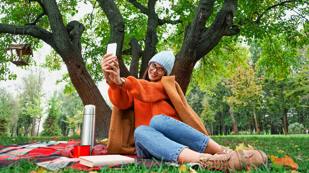 秋の公園でピクニック中にスマホで自撮りした幸せな若い女性 - 写真・画像