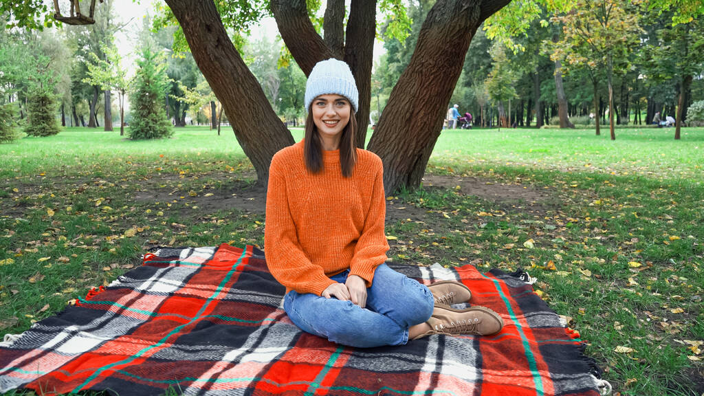 mulher elegante feliz sorrindo para a câmera enquanto sentado no cobertor xadrez no parque - Foto, Imagem