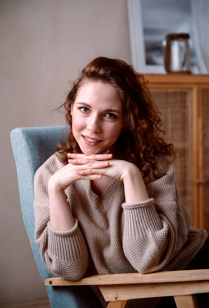 1 európai lány vörös hajjal bézs pulóverben ül egy székben és mosolyog, női portré - Fotó, kép