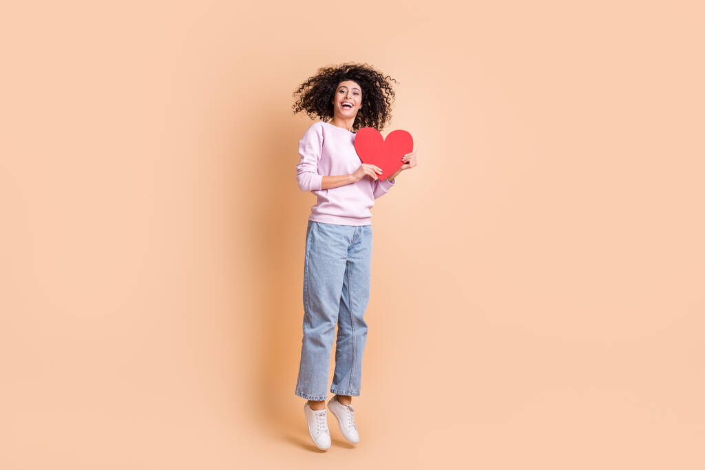 Pełny rozmiar zdjęcie optymistyczne kręcone dziewczyna skok z czerwonym sercem nosić sweter trampki izolowane na brzoskwiniowy kolor tła - Zdjęcie, obraz