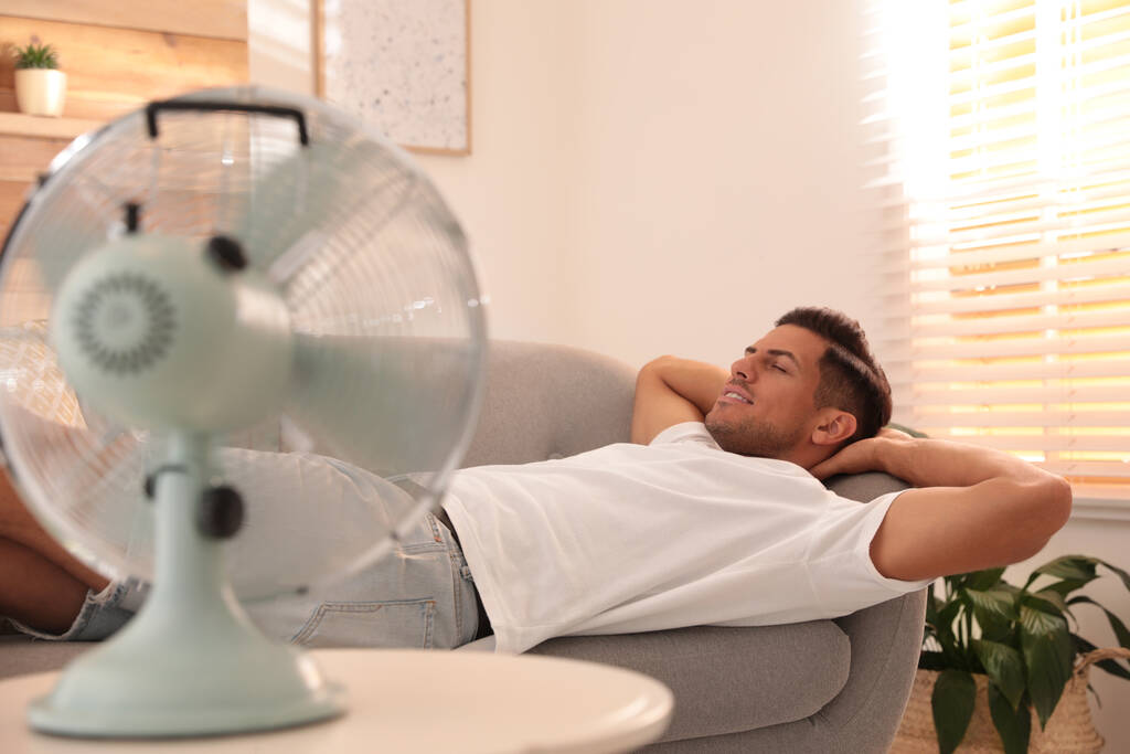 Человек наслаждается воздушным потоком от вентилятора на диване в гостиной. Летняя жара - Фото, изображение