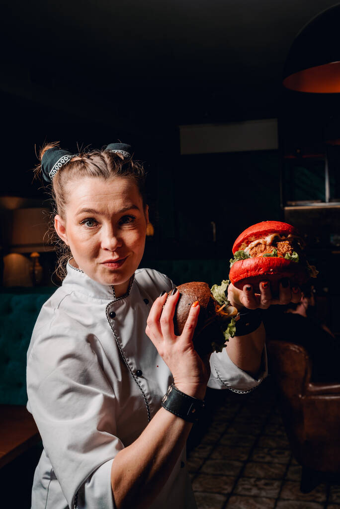 young woman chef with two burger in hands on dark background in cafe - Photo, Image