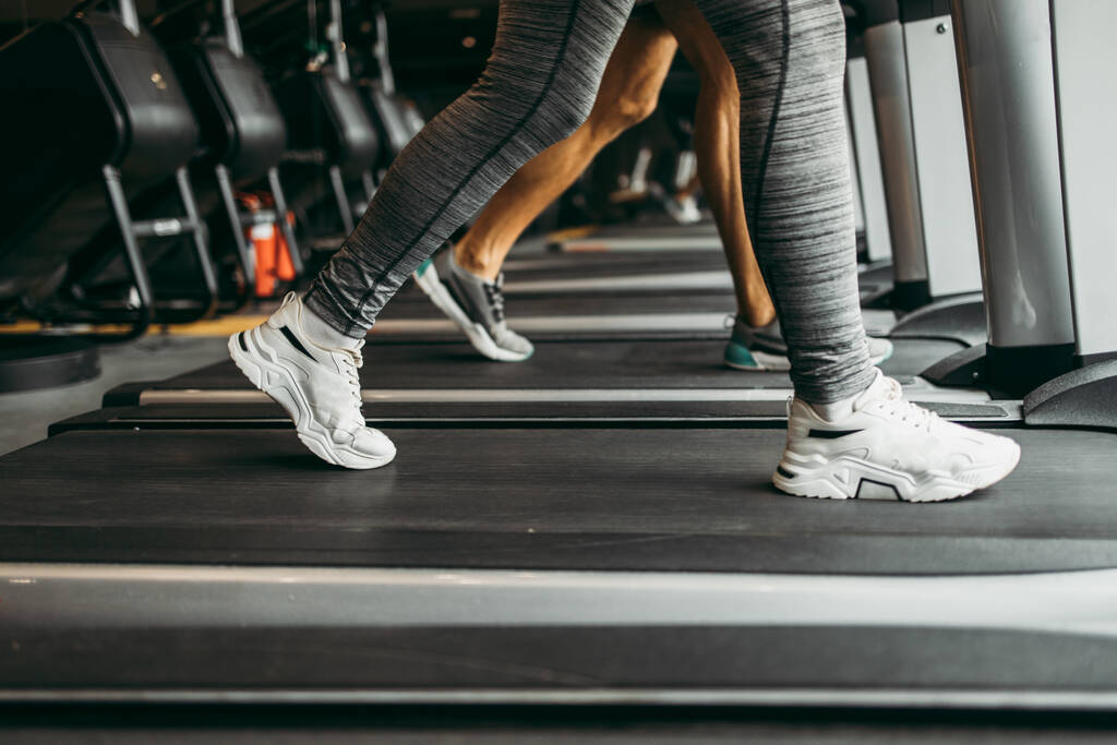 Молодая женщина и мужчина бегают по беговой дорожке в современном фитнес-зале. Закрыть рот на движущихся ногах. - Фото, изображение