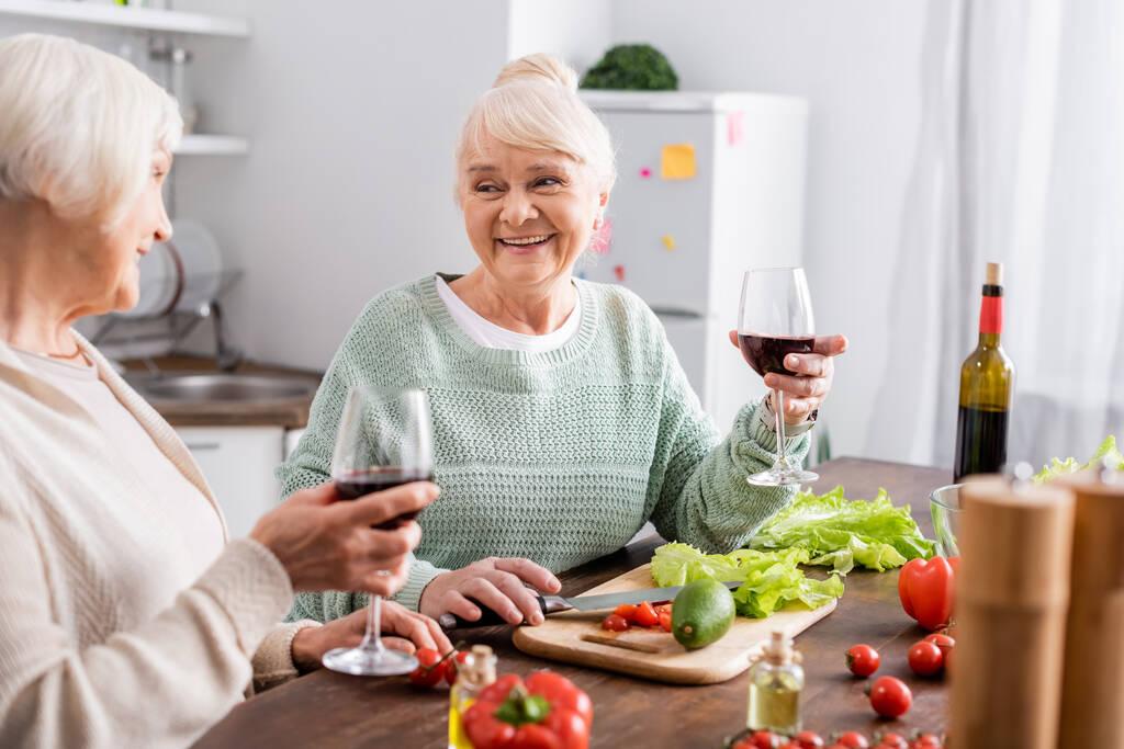 Gülümseyen yaşlı kadınlar masada taze sebzelerin yanında kırmızı şarap kadehleri tutarlar. - Fotoğraf, Görsel