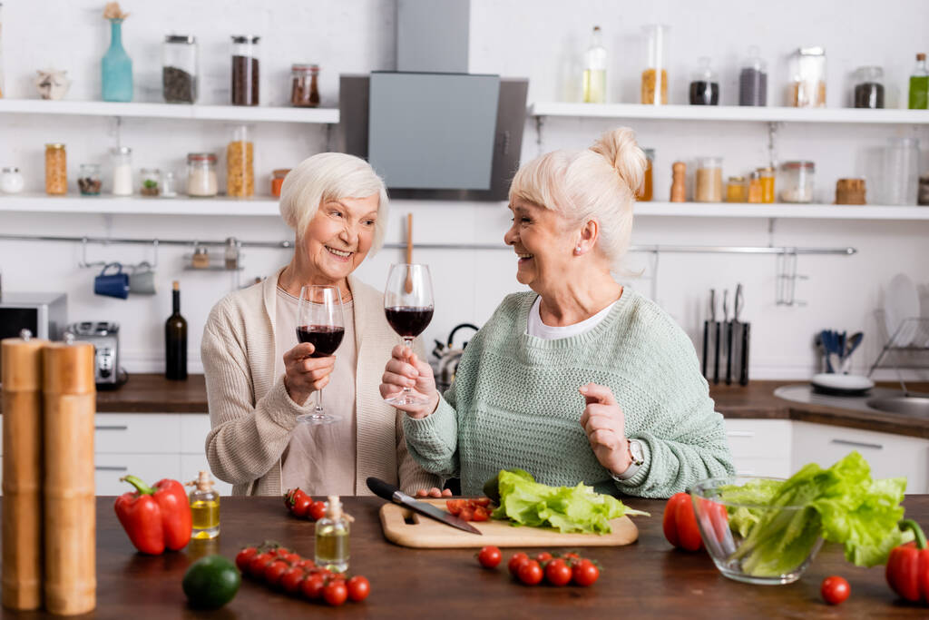 mujeres jubiladas sonrientes sosteniendo vasos de vino tinto y mirándose unos a otros cerca de verduras frescas en la mesa - Foto, imagen