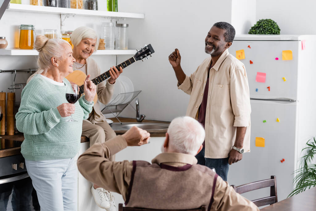 Glückliche Rentnerin spielt Akustikgitarre bei fröhlichen multikulturellen Freunden in der Küche  - Foto, Bild