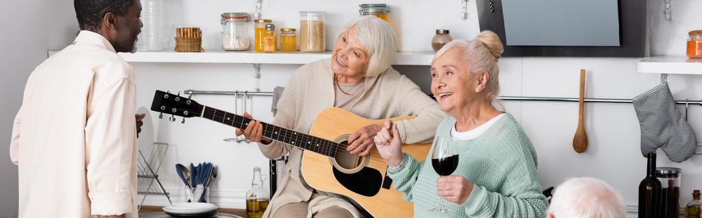 szczęśliwy emeryt kobieta gra na gitarze akustycznej w pobliżu starszych wielokulturowych przyjaciół w kuchni, baner - Zdjęcie, obraz