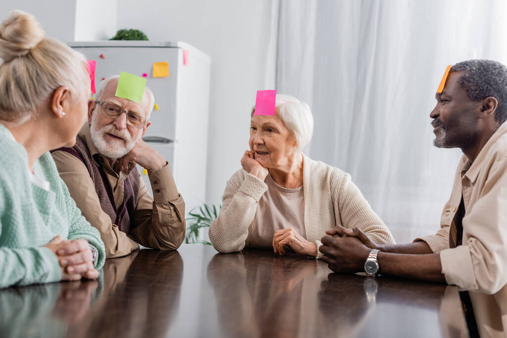 multiculturele senior vrienden met plakkerige notities op voorhoofd spelen spel in de keuken  - Foto, afbeelding