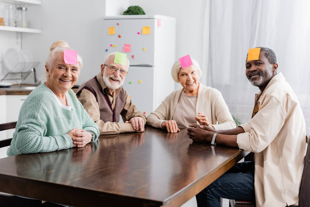 joyful multicultural senior friends with sticky notes on foreheads playing game in kitchen  - Photo, Image