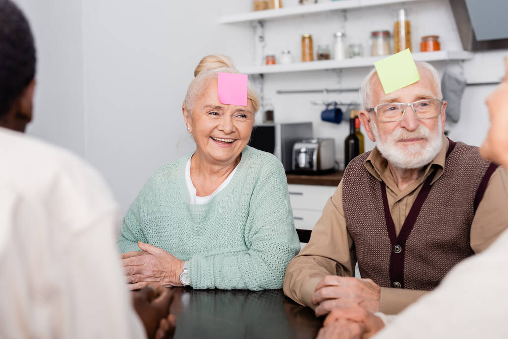 uomo e donna anziani sorridenti con note appiccicose colorate sulla fronte che giocano con amici multiculturali in primo piano sfocati - Foto, immagini