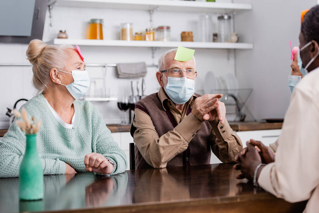 emeryci w maskach medycznych z lepkimi notatkami na czole grający w grę z wielokulturowymi przyjaciółmi - Zdjęcie, obraz