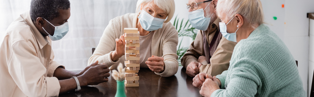personas jubiladas multiculturales en máscaras médicas jugando bloques de madera torre juego en casa, pancarta - Foto, imagen