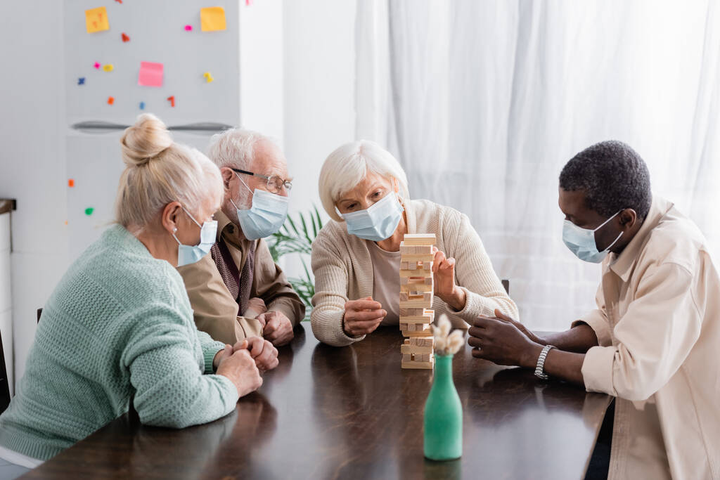 Multikulti-Freunde im Ruhestand in medizinischen Masken spielen zu Hause Turmholzklötze - Foto, Bild