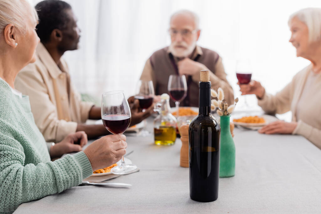 bottle of red wine near multicultural and senior friends having delicious lunch together on blurred background - Photo, Image