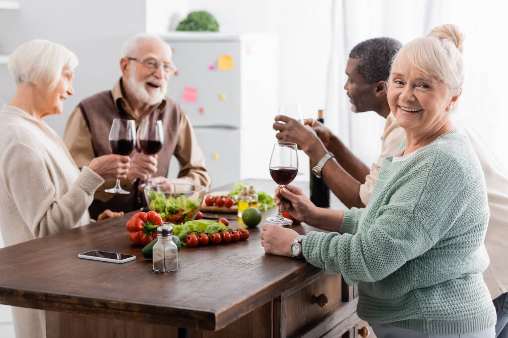 feliz anciana sosteniendo copa de vino tinto y sonriendo cerca de amigos jubilados multiculturales sobre fondo borroso - Foto, Imagen