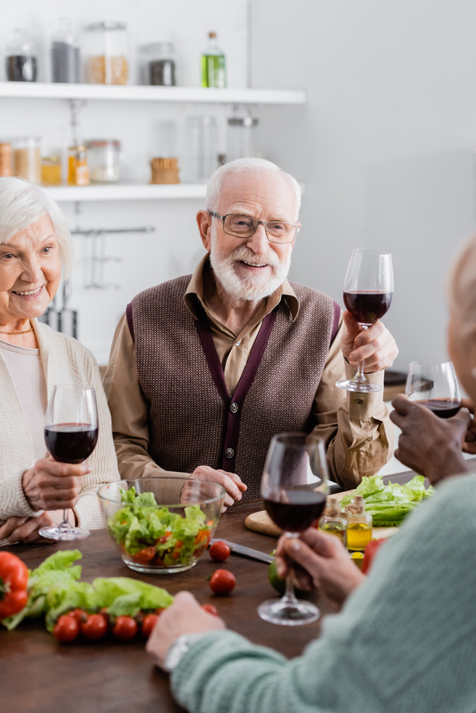 ανώτεροι διαφυλετικοί φίλοι κρατώντας ποτήρια με κρασί και χαμογελώντας κοντά σε λαχανικά στο τραπέζι - Φωτογραφία, εικόνα