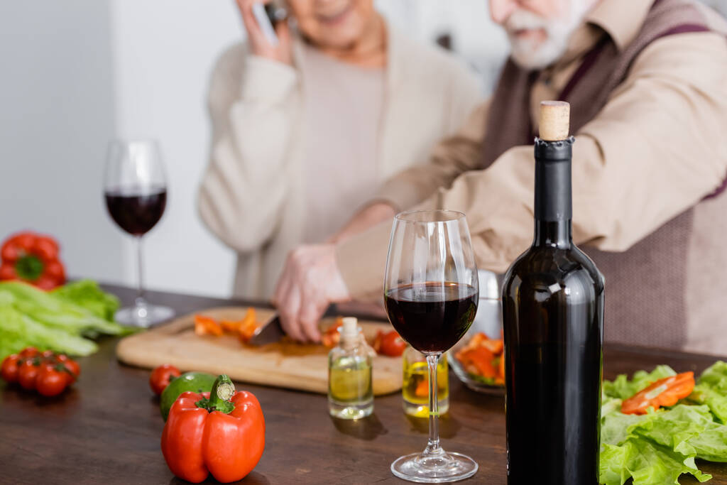 botella con vino tinto cerca de vidrio, verduras y pareja jubilada sobre fondo borroso  - Foto, imagen
