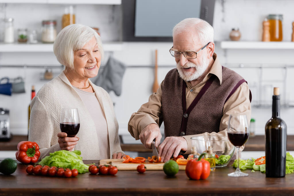 щасливий старший чоловік ріже овочі біля усміхненої дружини з келихом вина
 - Фото, зображення