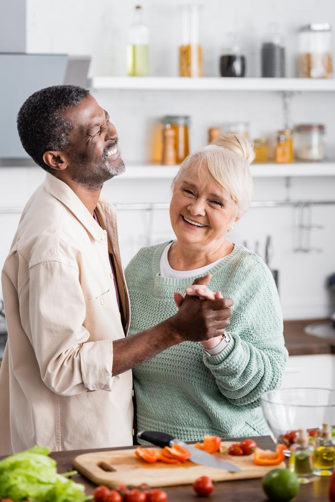 senior multicultural husband and wife smiling while holding hands in kitchen  - Photo, Image