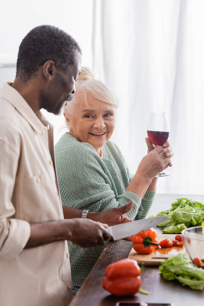 Uomo afroamericano taglio peperone vicino moglie anziana con bicchiere di vino - Foto, immagini