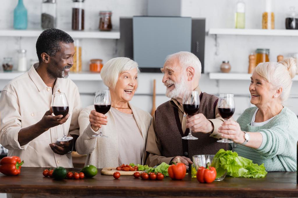 pensionati multiculturali gioiosi che tengono bicchieri su vino vicino a verdure a tavola - Foto, immagini