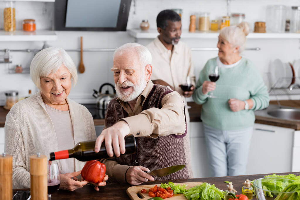 glücklicher älterer Mann gießt Wein in Glas neben Rentnerin und multikulturellen Freunden auf verschwommenem Hintergrund  - Foto, Bild
