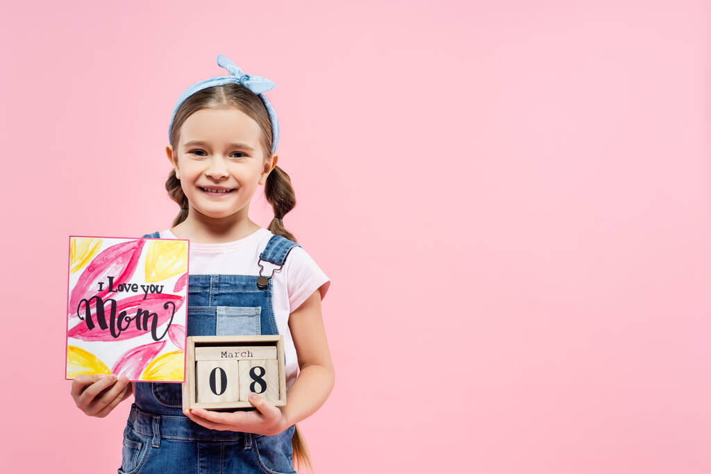 счастливый малыш с поздравительной открыткой "Я люблю тебя, мама" и деревянными кубиками с 8 Марта на розовом фоне  - Фото, изображение