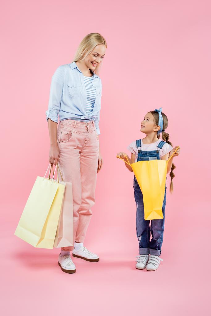 full length of cheerful mother and daughter looking at each other while holding shopping bags on pink - Photo, Image